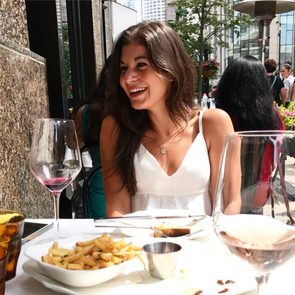 A woman smiles while dining outdoors, surrounded by wine glasses and plates of food, with an urban setting in the background.