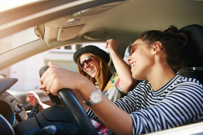 Two trendy attractive young woman singing along to the music as they drive along in the car through town viewed through the open side window