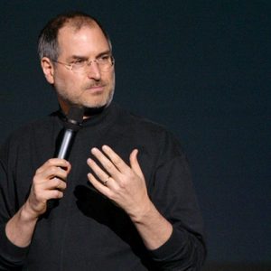 A person holds a microphone while speaking, set against a dark background, wearing glasses and a black turtleneck.