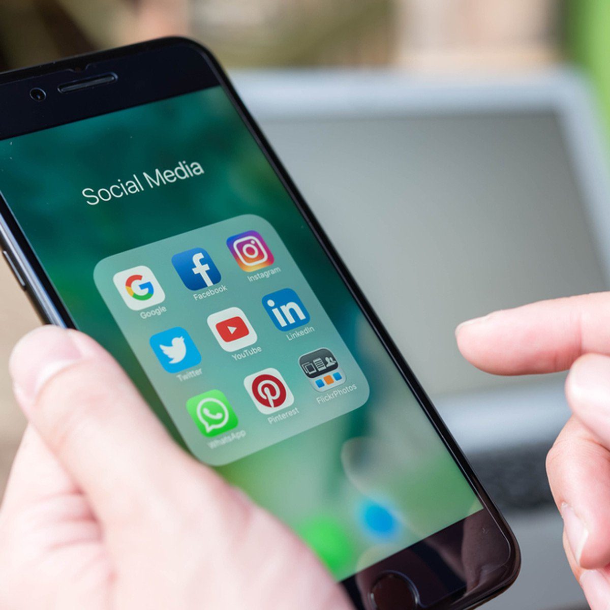 Smartphone displaying social media apps, held by a hand; finger pointing toward screen, with a blurred laptop in the background.