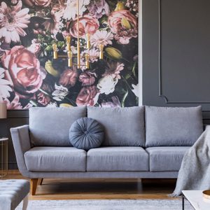 Blanket on grey couch in living room interior with flowers wallpaper and lamp on table.