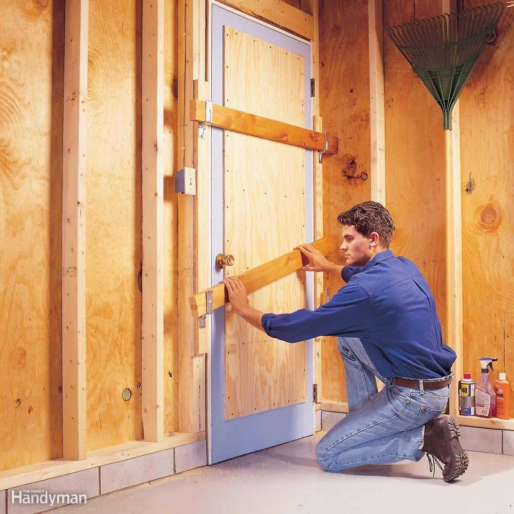 Person securing door with wooden bars, kneeling in unfinished wooden room; various tools and supplies on the floor. "Handyman" text in lower left corner.