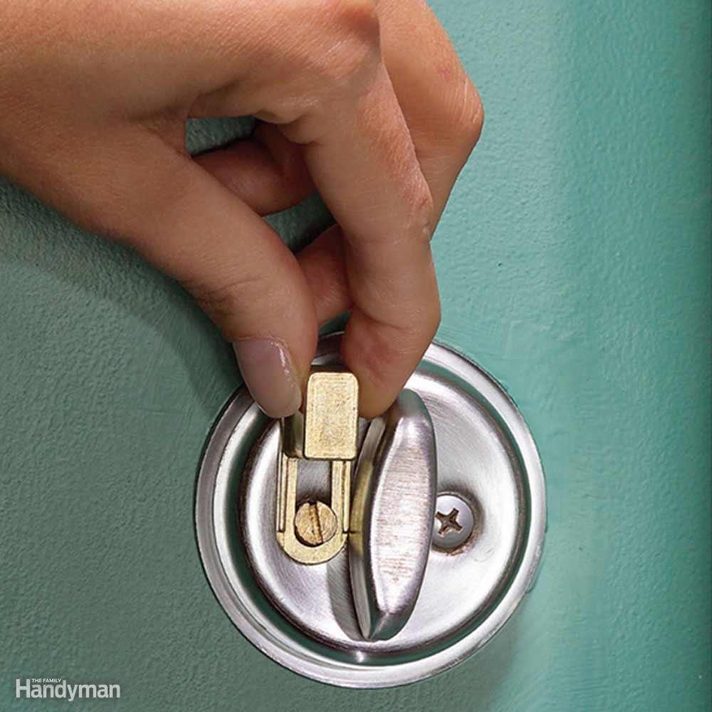 Hand adjusts a brass slider on a silver deadbolt, against a teal door. Small "The Family Handyman" logo in the bottom corner.