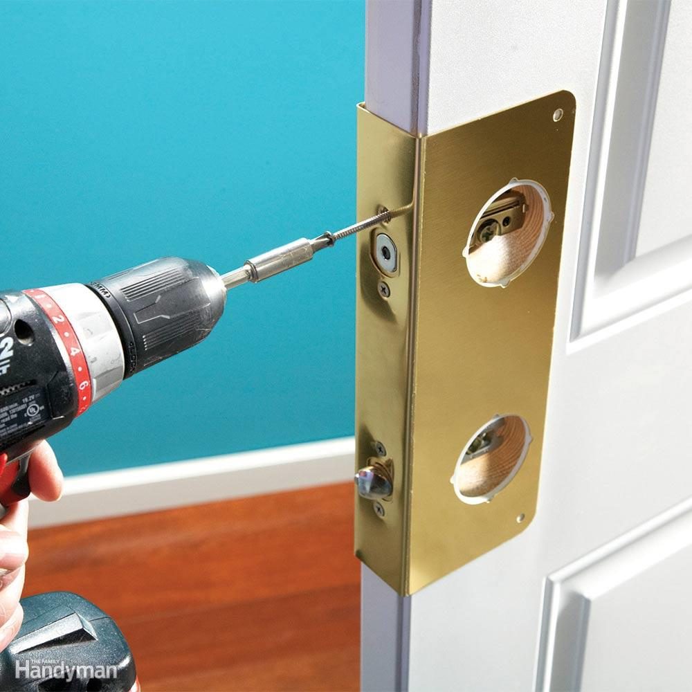 Drill installs screws into a gold door security plate; door is ajar in a room with blue walls and wooden floor.