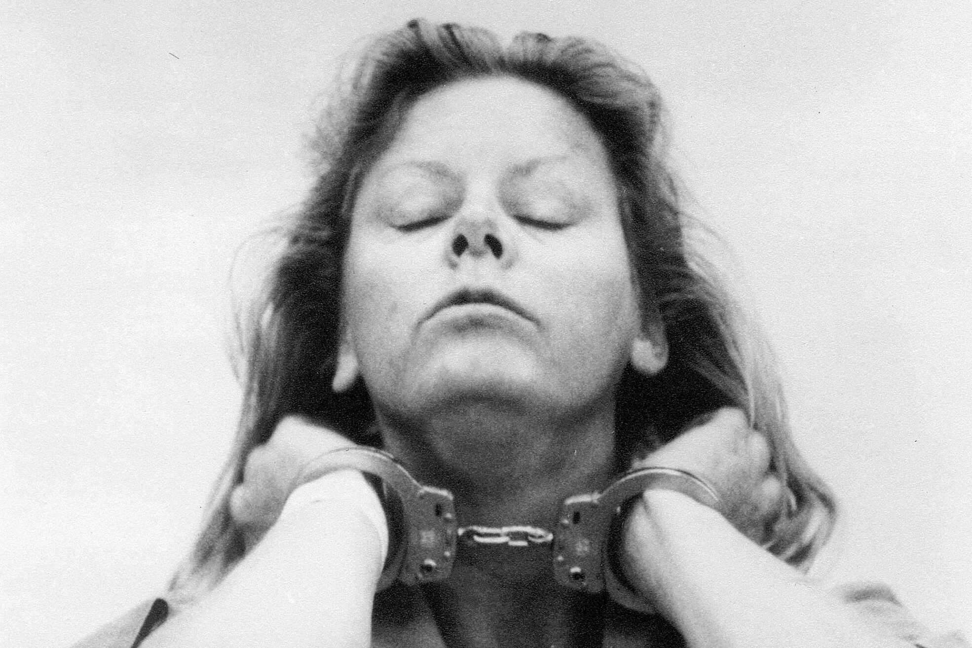 Person stands with handcuffs, eyes closed, in a jumpsuit against a plain background.