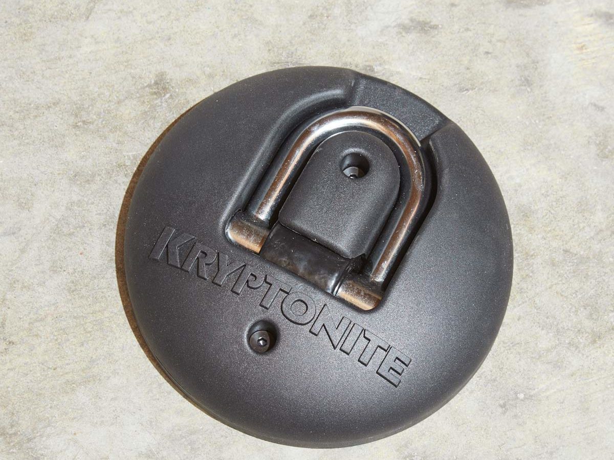 Black, round Kryptonite lock anchor mounted on a concrete surface, featuring a metal U-shaped shackle in the center.