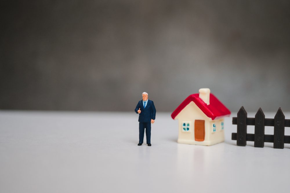 A small figurine in a suit gives a thumbs-up next to a toy house with a red roof and a black fence on a plain surface.
