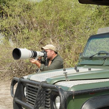 Photographer uses a large telephoto lens while sitting on a green safari vehicle, surrounded by bushes and open landscape.