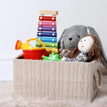 A knitted basket holds toys, including a xylophone, watering can, doll, and stuffed animal, on a fluffy rug beside a white wall.
