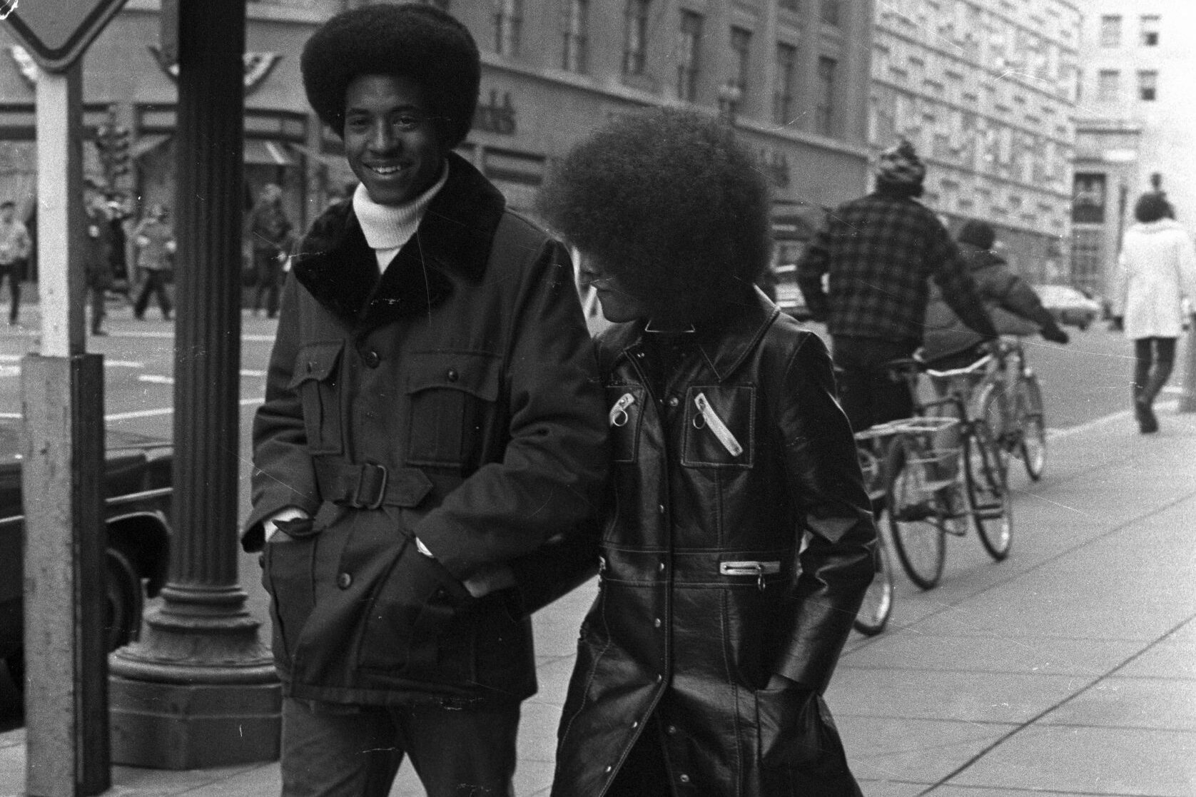 Two people walk on a city sidewalk, wearing stylish 1970s outfits, with a street sign and bicycles in the background.