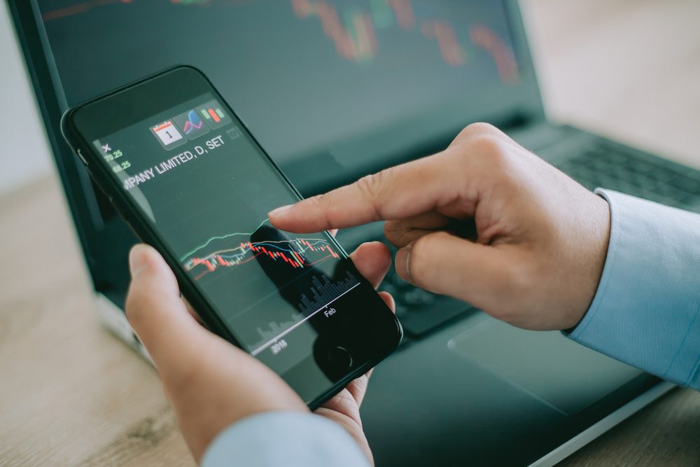 Business man point to the smart phone show financial market chart and Blurred background of a laptop show financial market chart, Stock market concept.