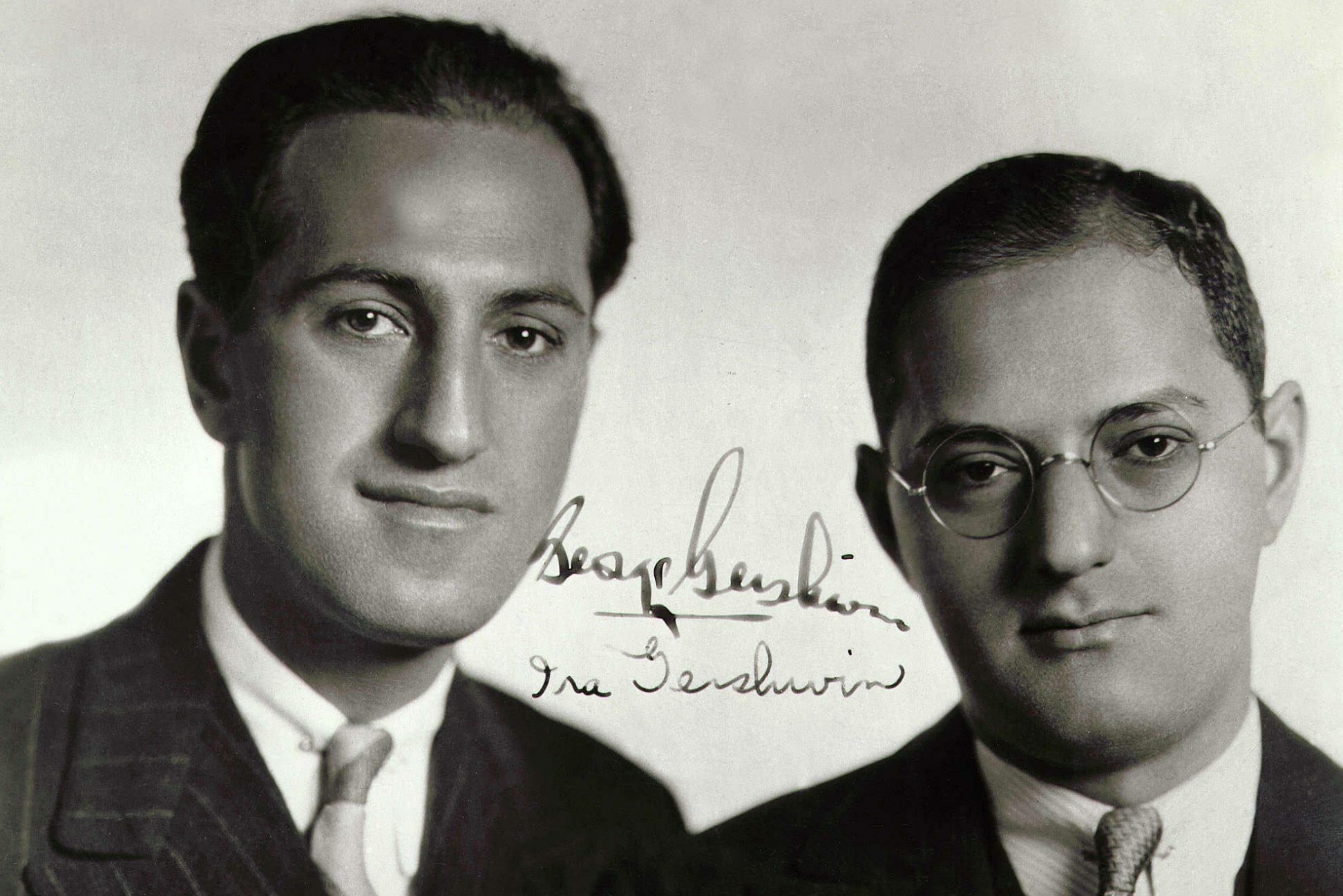 Two men sit closely together, wearing suits and ties, in a formal, monochrome setting. The image is signed "George Gershwin" and "Ira Gershwin."