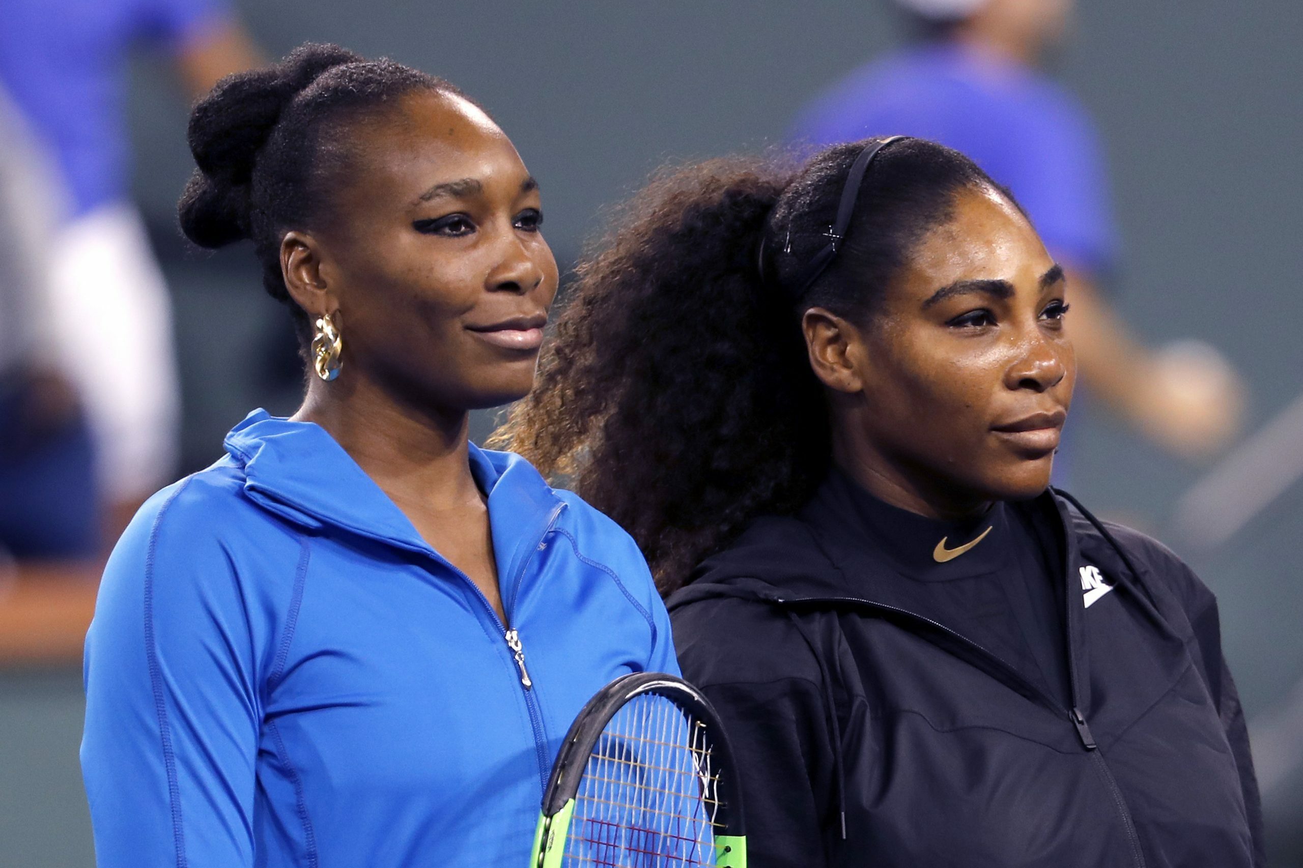 Two women stand together, one holding a tennis racket, on a sports court, wearing blue and black athletic jackets.