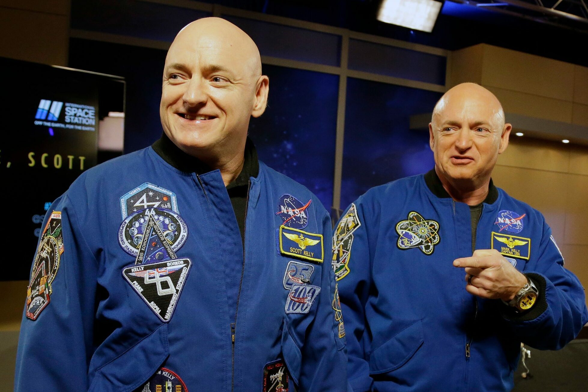 Two astronauts in blue jackets stand smiling; one points at the other's patches. A screen displays "WELCOME HOME, SCOTT" in a room.