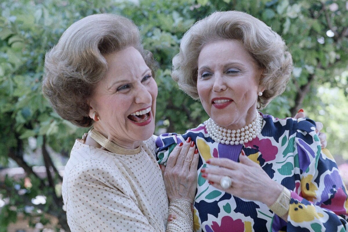 Two women laughing, one in a polka-dot blouse, the other in a colorful floral dress with pearls, standing outdoors among green trees.