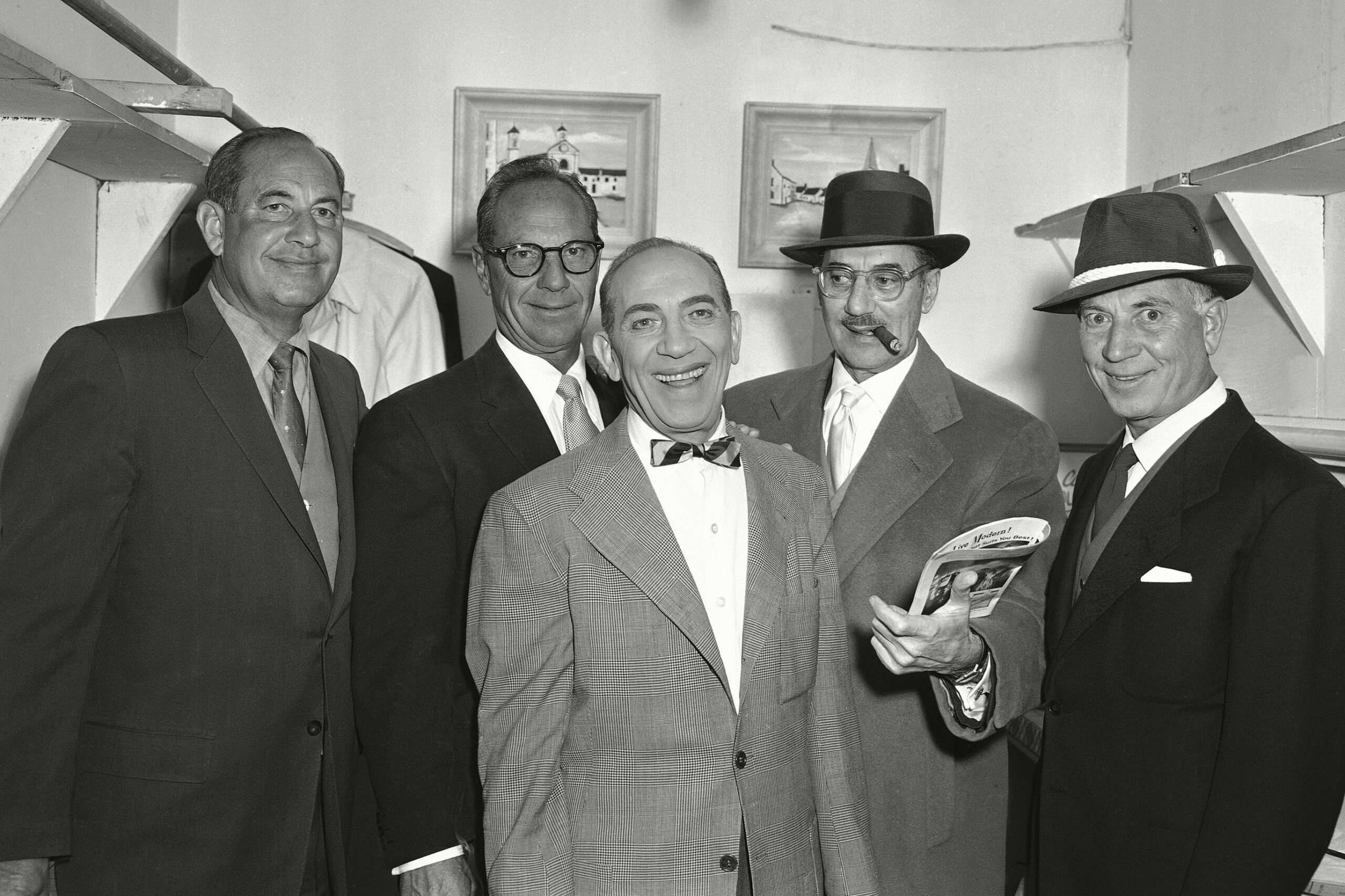 Five men stand smiling in suits and hats, one holding a magazine, inside a room with paintings on the walls.