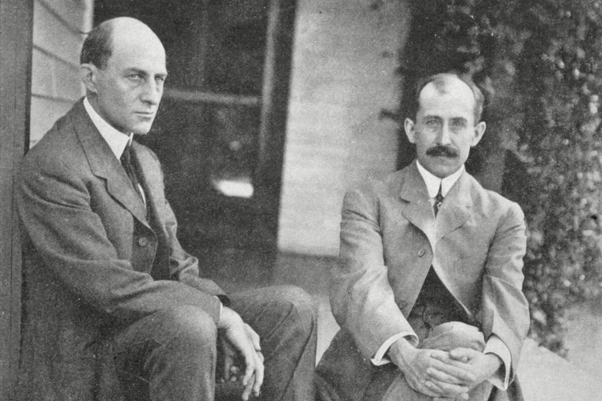 Two men sitting on wooden steps, both in suits, with one crossing his legs. The background shows a building with plants.
