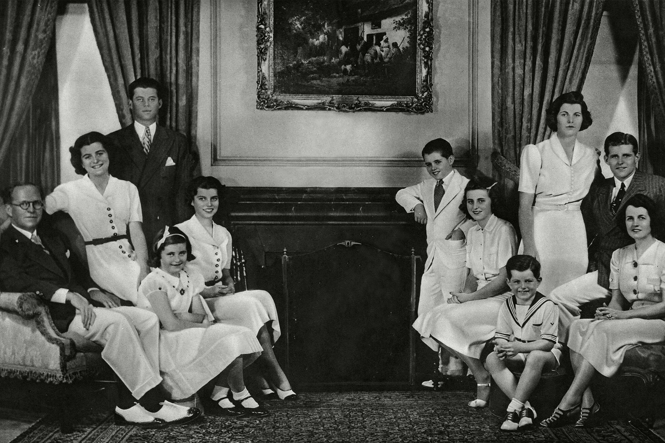 Family poses together in formal attire, seated and standing around a grand room with a framed painting and draped curtains in the background.