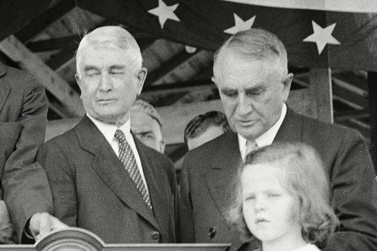 Two men stand solemnly beside a child, in front of a starry banner, during a formal event.