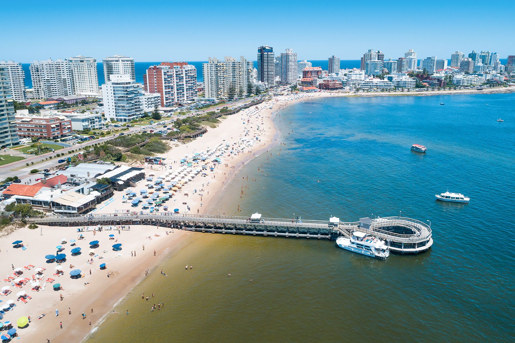 Beach with sunbathers and umbrellas, a pier extends into the sea with moored boats, city skyline and buildings in the background.