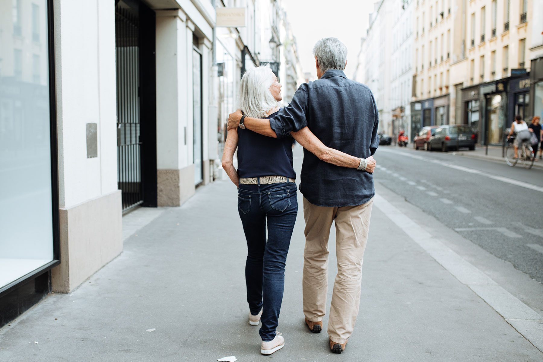 An elderly couple walks arm-in-arm down a city street, surrounded by tall buildings and a few people in the distance.