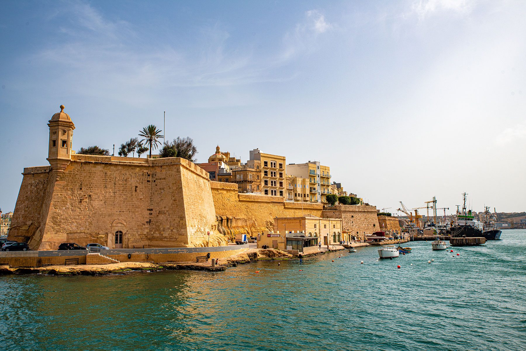 Fortress stands prominently, overlooking a harbor. Boats float on calm water, and stone buildings and trees line the shoreline under a clear sky.