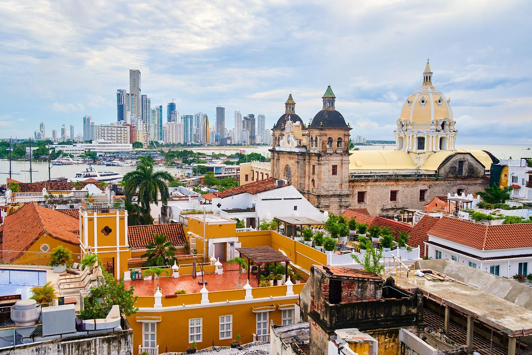 Colorful rooftops surround a historic church, framed by modern skyscrapers under a partly cloudy sky in a coastal city.