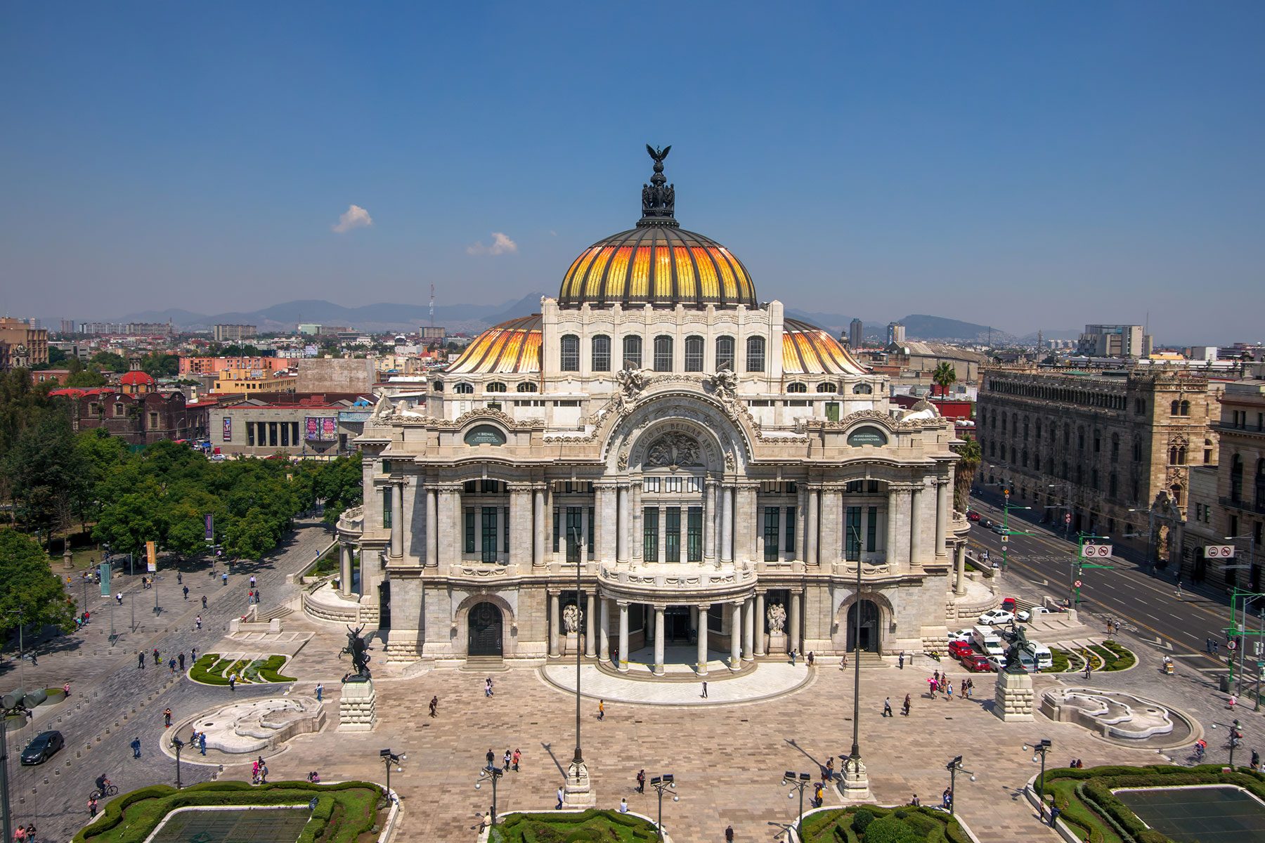 Grand domed building dominates plaza, featuring sculptural elements and stained glass, surrounded by bustling urban street with pedestrians and parked cars, under clear blue sky.