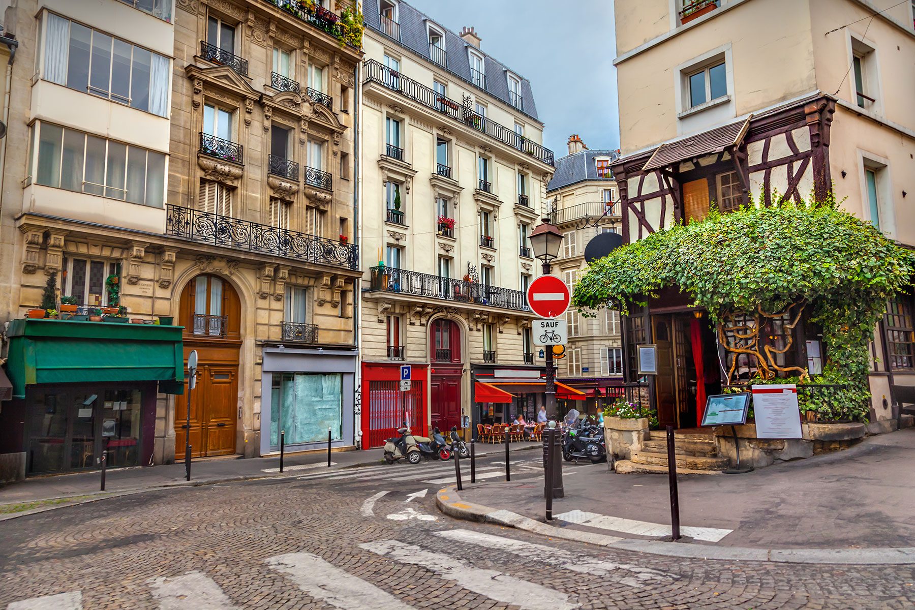 Buildings line a cobblestone street; red awnings cover a cafÃ©. A vine-covered structure and a "no entry" sign with "SAUF" fill the scene.