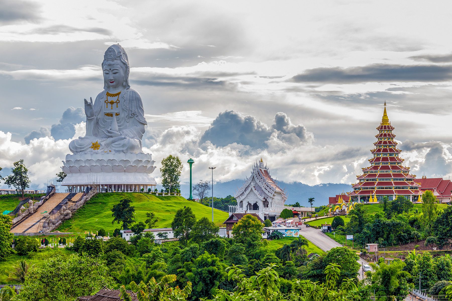 A large white statue sits peacefully on a green hilltop, with a decorative temple and cloudy sky in the background.