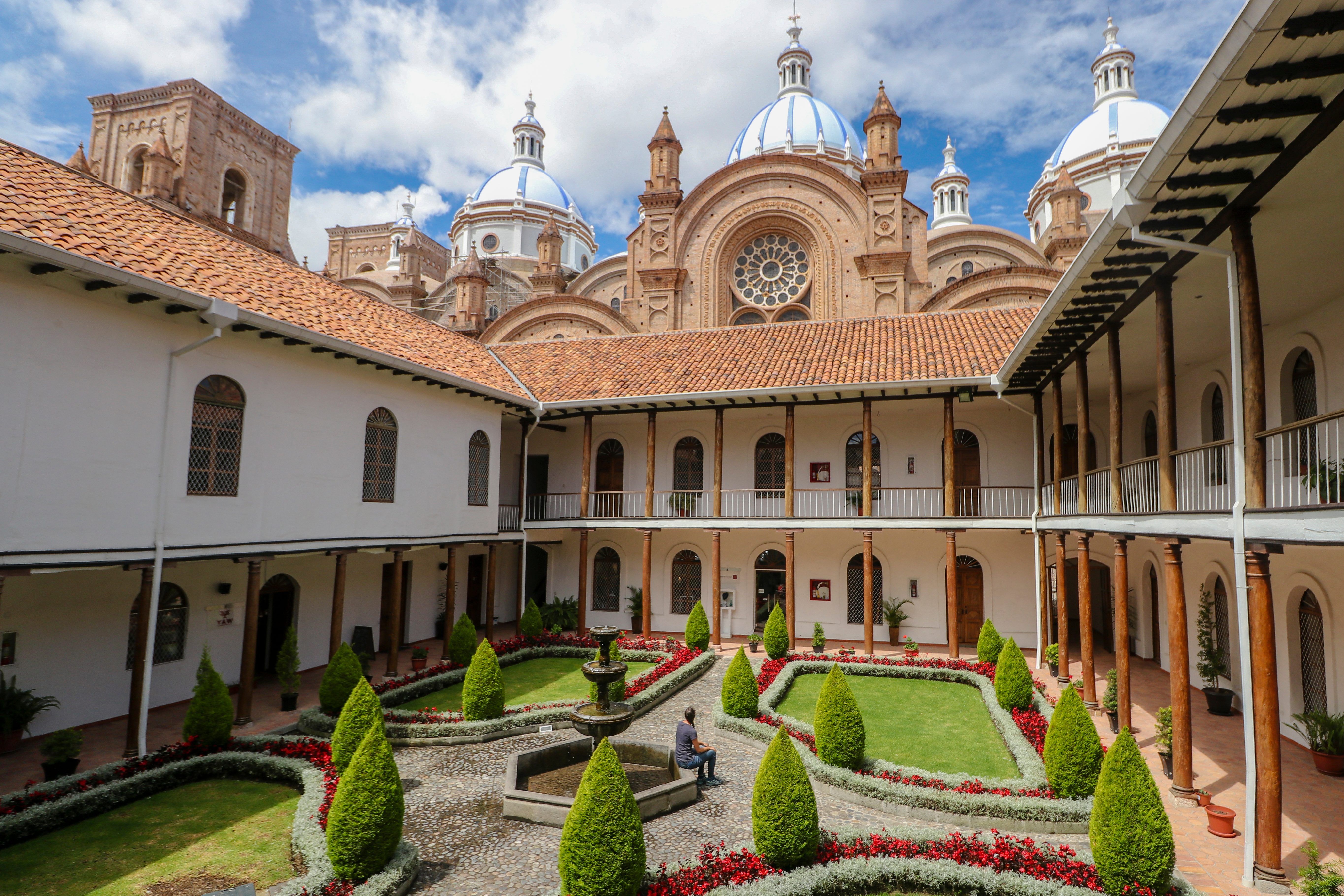 Fountain sits in a courtyard garden, surrounded by manicured bushes and a two-story colonial building, with a cathedral featuring domes in the background.