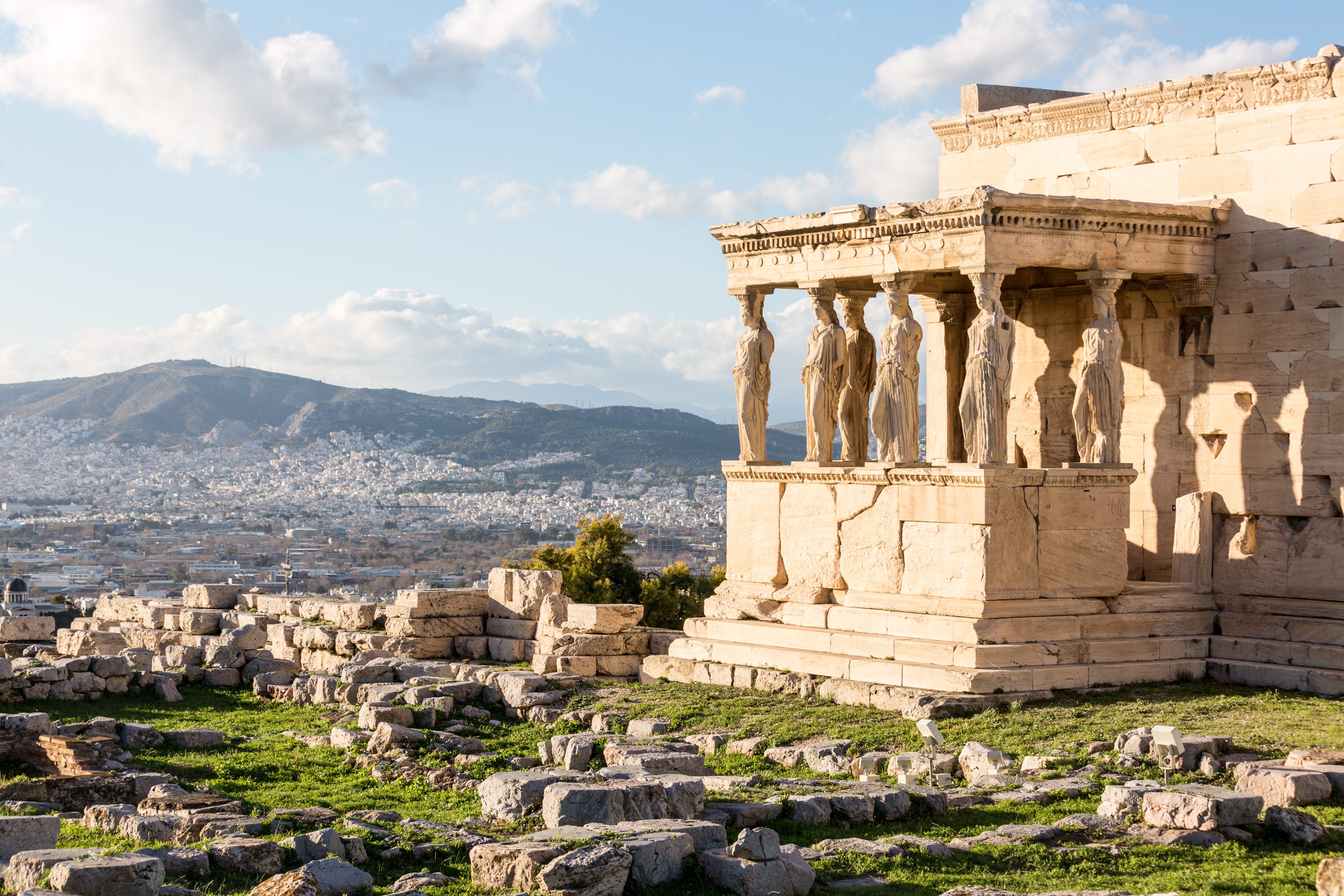 Ancient stone statues stand on a temple porch, overlooking a cityscape framed by distant mountains and scattered ruins in a sunlit landscape.
