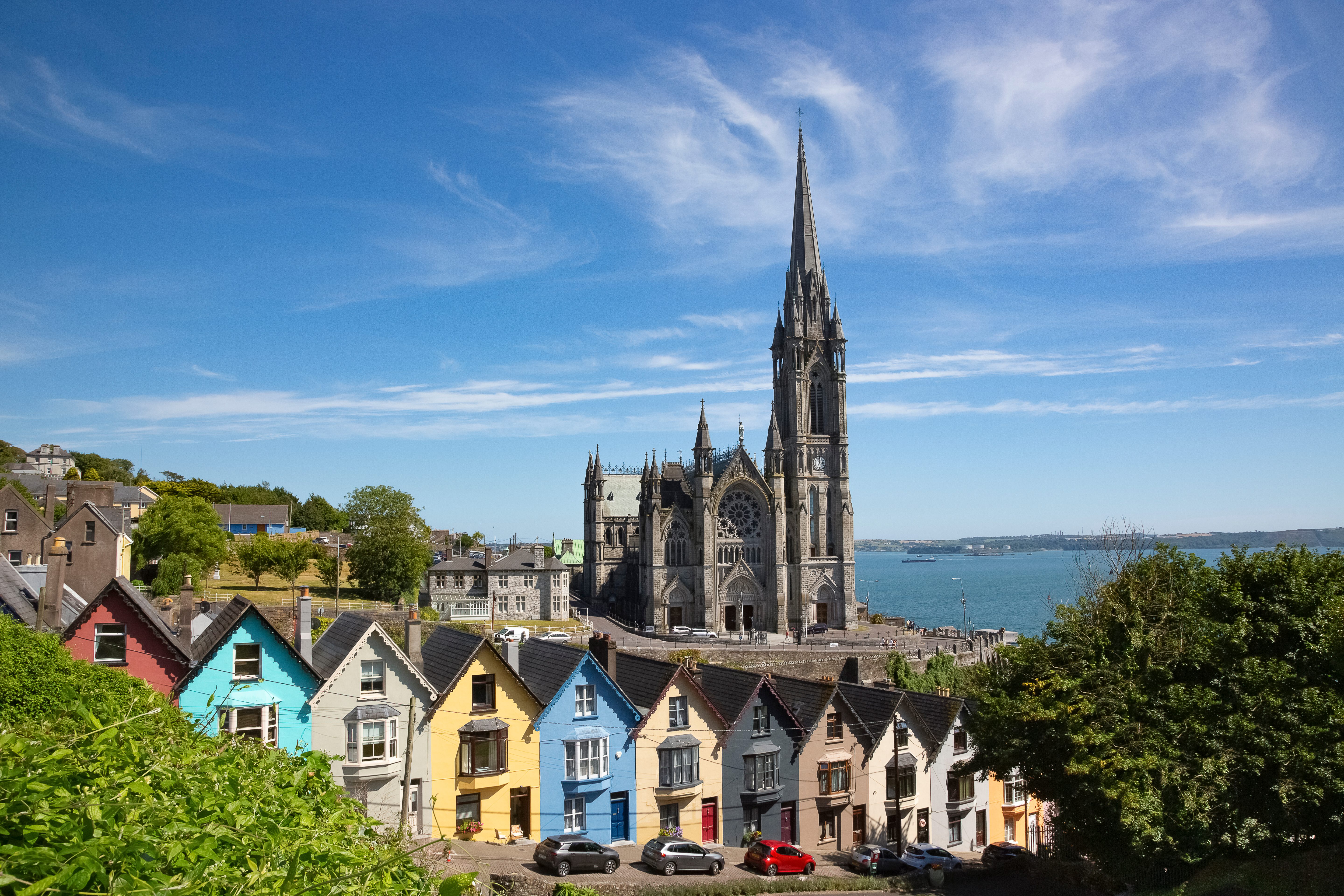 Colorful houses line a hillside, facing a towering cathedral with a pointed spire, against a backdrop of blue sky and distant water.