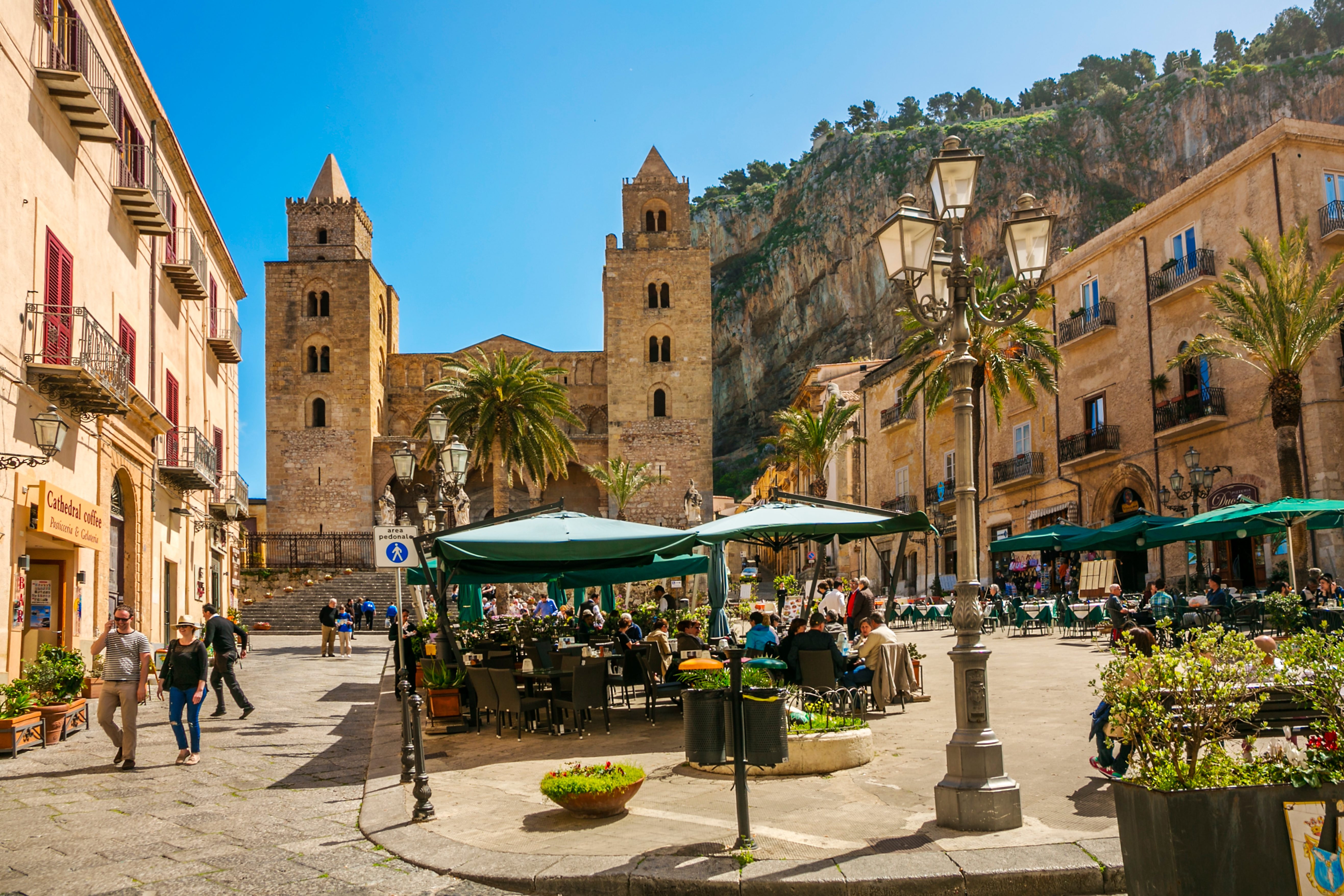 Cafe patrons sit under green umbrellas with cathedral towers in the background, surrounded by historic buildings and a mountainous landscape.