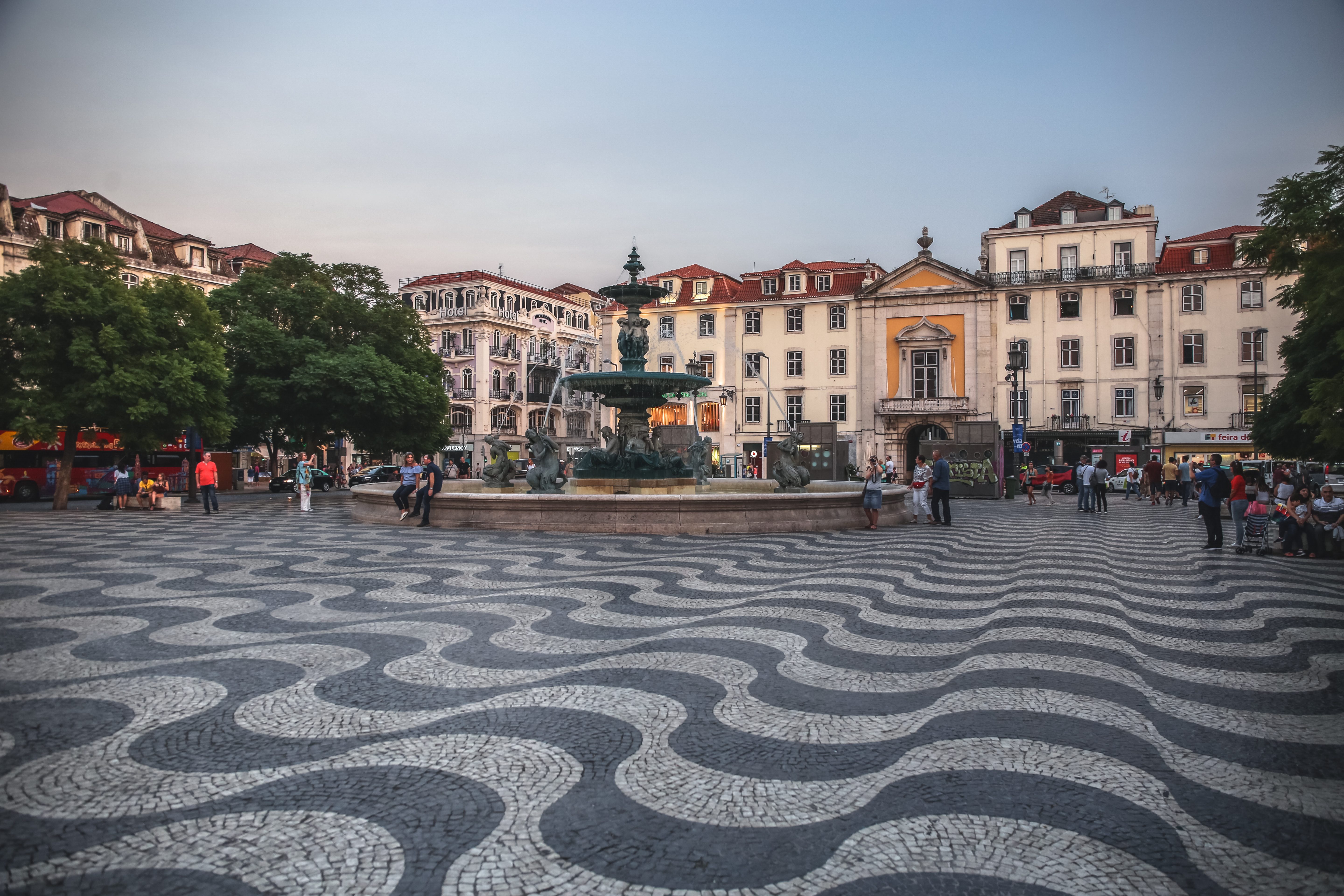 Fountain sits in a vibrant square, surrounded by historic buildings and people walking on a patterned cobblestone ground.