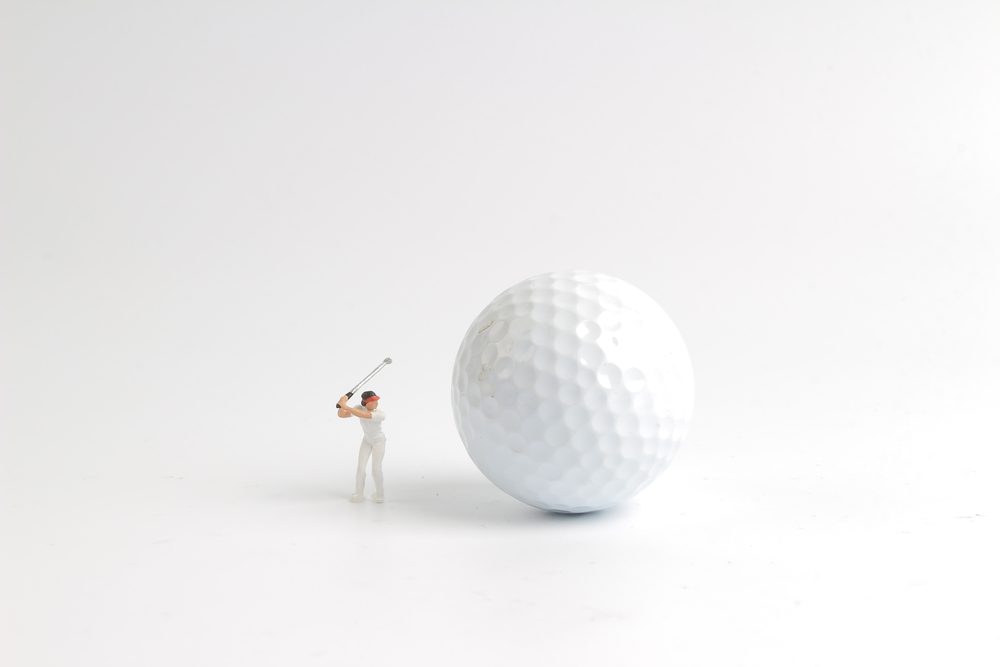 A tiny golfer swings beside an oversized golf ball on a plain white background.