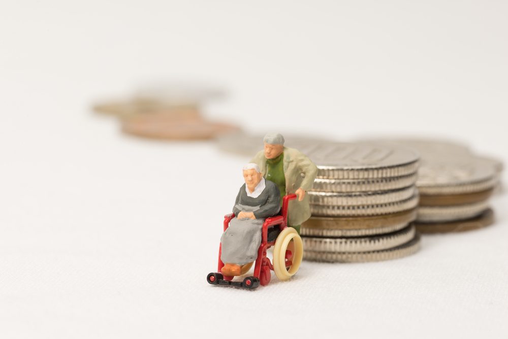 Miniature figures: a person pushes another in a wheelchair beside stacks of coins, symbolizing financial planning and elderly care.