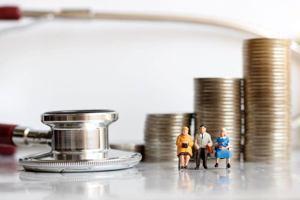 Miniature figures sit on a stethoscope beside stacks of coins, symbolizing healthcare costs and financial planning.