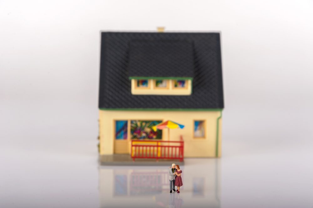 Small figurines embrace in front of a model house with a dark roof, colorful windows, and a yellow umbrella on its porch.