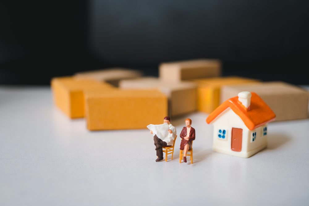 Two miniature figures sit reading by a small toy house, surrounded by yellow and brown blocks in a simple setting.