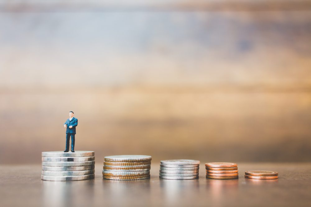 A small figure stands on a pile of coins, with descending stacks beside, against a blurred wooden background.