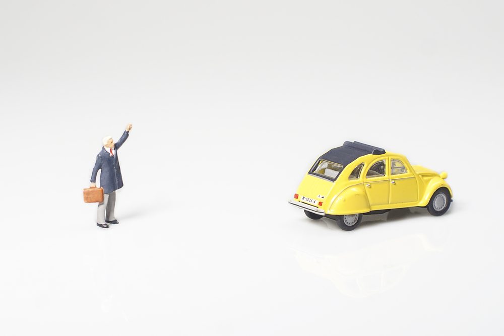 Miniature figure waves at a yellow toy car on a smooth, white surface.