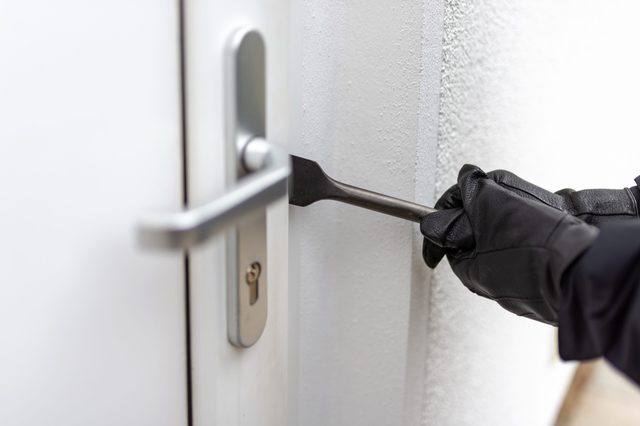 Gloved hand pries open door with crowbar, in a close-up view against a plain, light-colored wall.