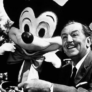 A person and large cartoon character smile together in front of a castle, with trees in the background.