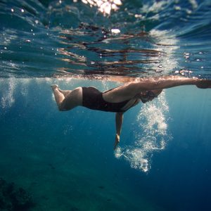 Person swimming underwater, wearing a black swimsuit, in clear blue ocean water, creating bubbles as they move forward.
