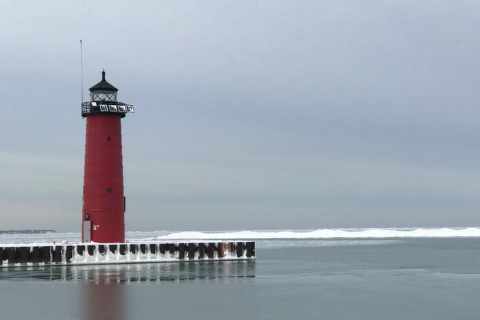Red lighthouse stands silently, surrounded by icy waters and a cloudy sky, on a snow-dusted pier.