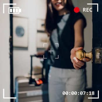 A person opens a door while holding a suitcase handle, recorded by a video camera in a room with framed pictures.
