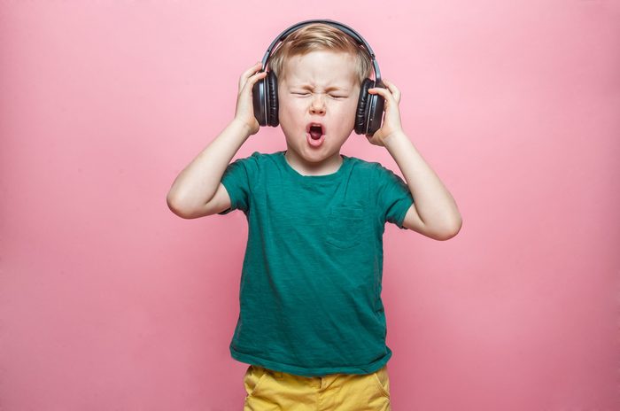 Stylish teen boy listening music in headphones and singing against pink background. School child listening loud music in wireless earphones and dancing.