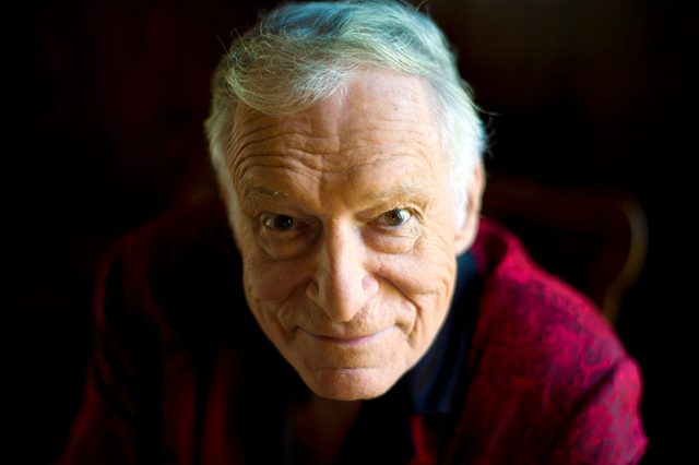 An elderly man with white hair smiles slightly, wearing a red robe, in a dimly lit setting.