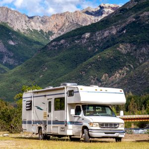 Camper parked in a grassy area, surrounded by trees, with mountains in the background and a river nearby.